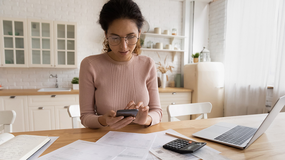 woman sitting at a table holding a smartphone surrounded by paperwork and a laptop